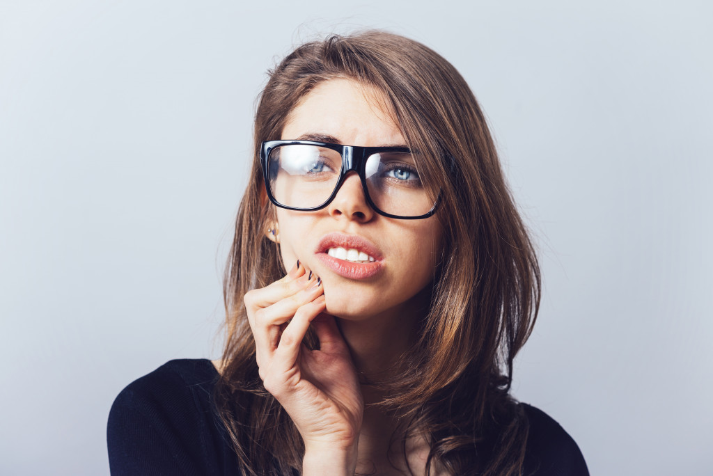 Young woman wearing glasses looks in pain while holding her teeth 