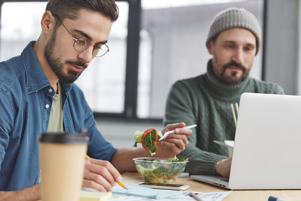 Concentrated bearded stylish male office worker being vegan, looks attentively at documents and eats fresh vegetable salad and his experienced fashionable colleague keyboards on laptop computer.