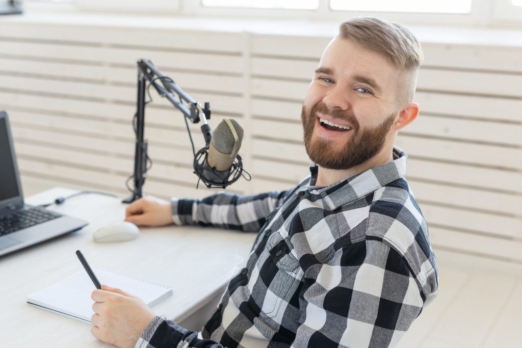 A happy man sitting in front of a laptop and microphone to make a podcast
